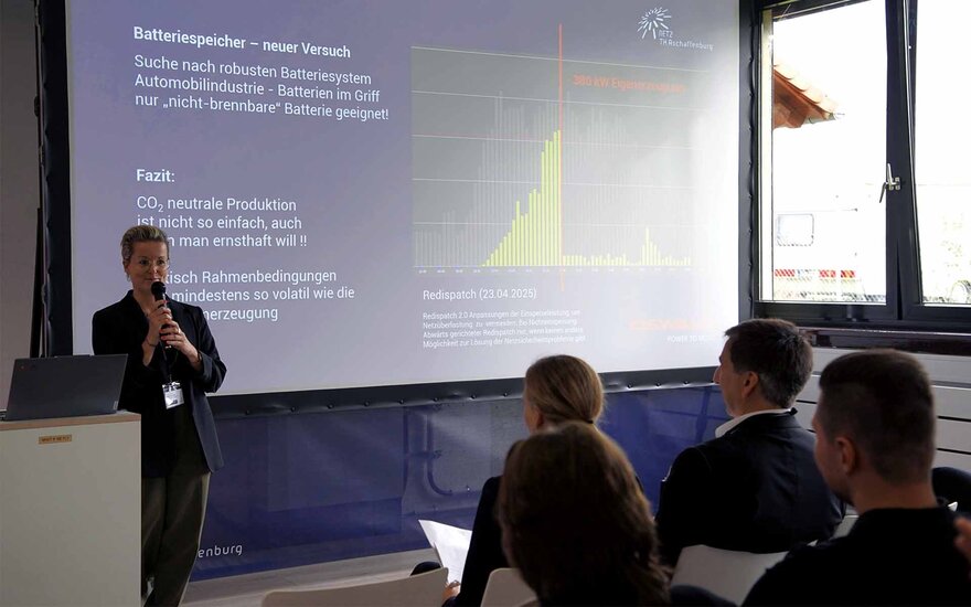 Eine Frau im Blazer hält einen Vortrag. Auf der Folie ist ein Diagramm zum Thema CO2-neutraler Produktion von Batteriesystemen. 