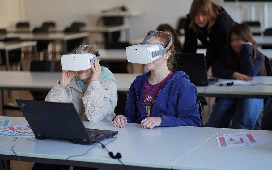 Zwei Mädchen sitzen mit VR-Brillen vor einem aufgeklappten Laptop.