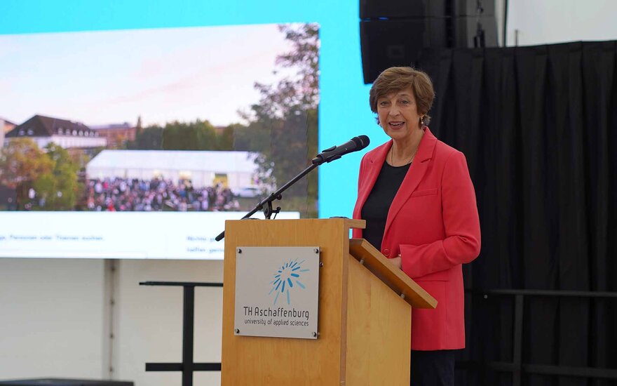 Frau mit braunen kurzen Haaren und rotem Blazer am Rednerpult vor Leinwand