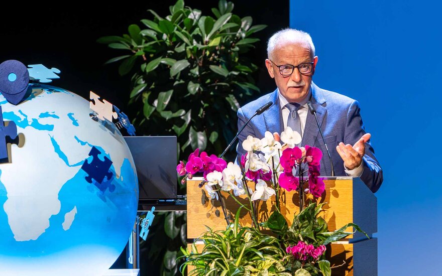 Älterer Herr mit Glatze und Brille im hellblauen Anzug am blumengeschmückten Rednerpult