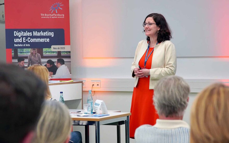 Professorin Bertels spricht im roten Kleid neben rot-blauem Studiengangsrollup zu den Gästen