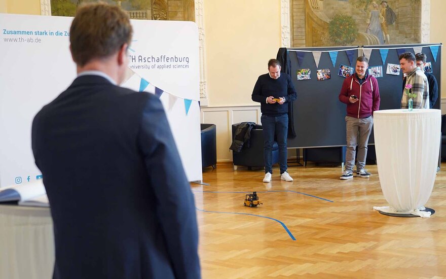Timm steuert mit Fernbedienung einen Mini-Gabelstapler, Manuel und Luca stehen neben ihm, Prof. Zindler im Vordergrund