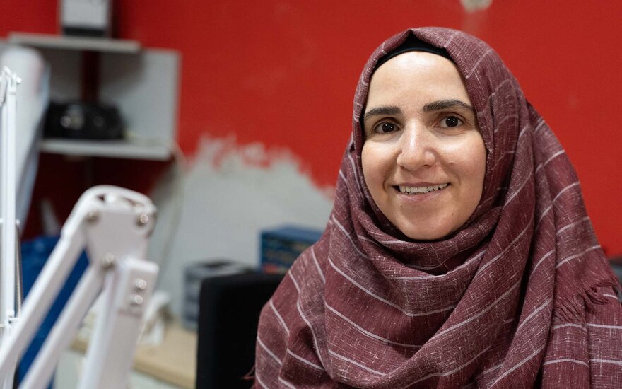 Portraitaufnahme von Amina in der sie lächelt. Im unscharfen Hintergrund, erkennt man die rote Wand der Nähwerkstatt, eine Nähmaschine und ein weißes Regal.