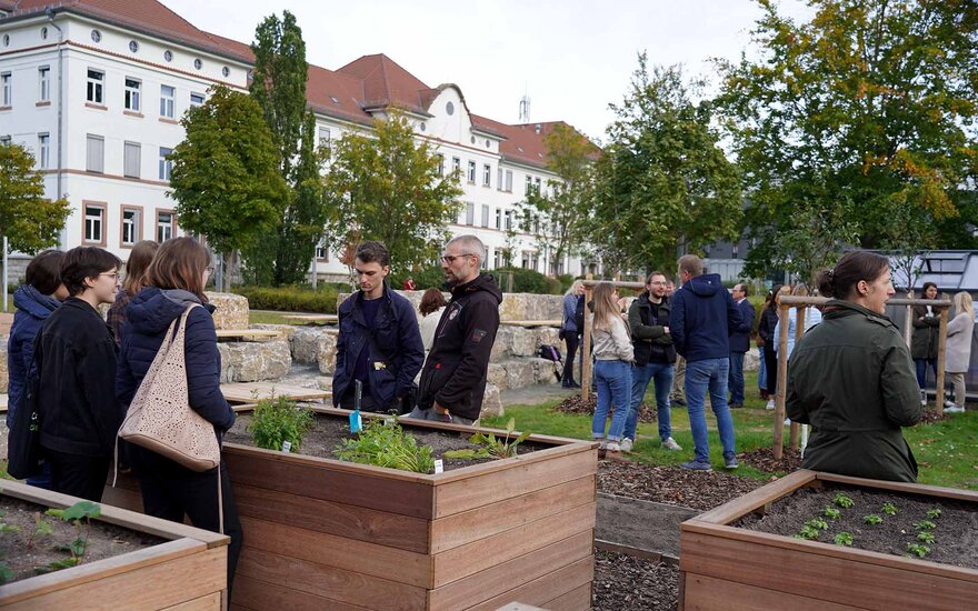 Viele Hochschulangehörige stehen um die Hochbeete herum und an Stehtischen und schauen sich den neuen Garten an.