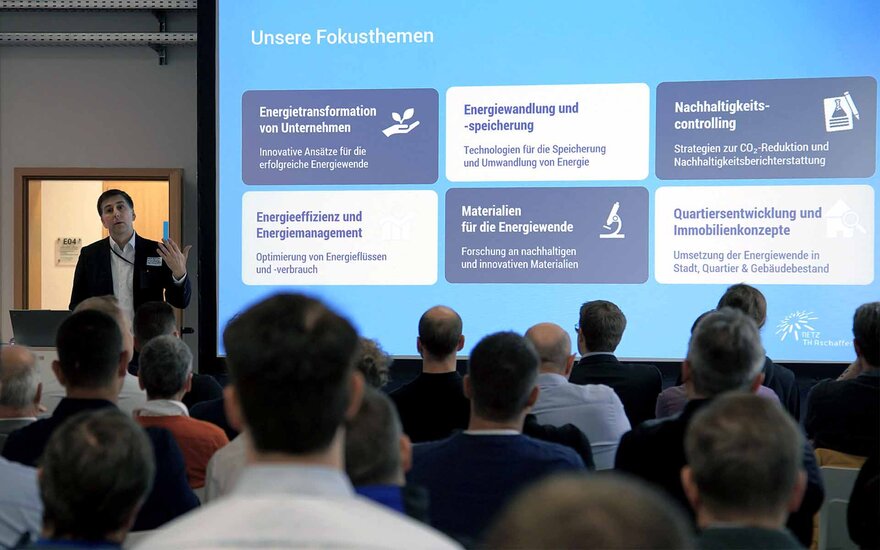 Mann steht neben einer Bildschirmpräsentation. Er spricht zu einem Publikum.