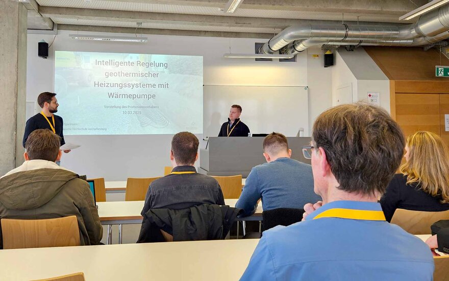 Ein Doktorand steht mit dem Moderator vor einer Leinwand, im Vordergrund sitzen Zuhörende