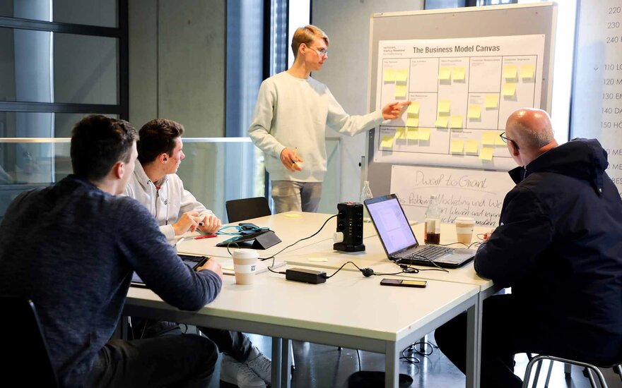 Ein junger Mann klebt ein Post-it an eine Flip Chart, drei weiter Männer schauen im am Tisch sitzend zu