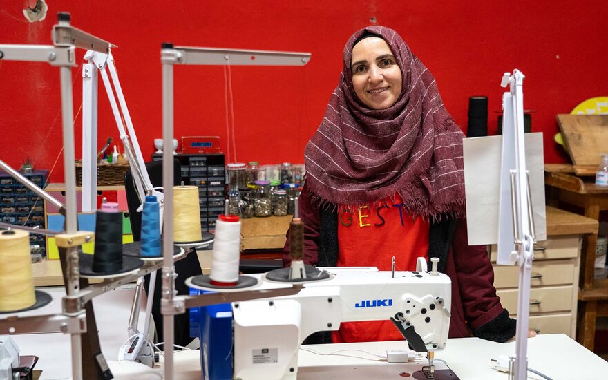Amina steht vor einer Werkbank in der Nähwerkstatt von Gesta e.V. und lächelt in die Kamera. Sie trägt ein rotes T-Shirt, das mit dem Wort "Gesta" bunt bestickt ist. Vor ihr befindet sich eine Nähmaschine, unterschiedlich gefärbte Nähfäden, die Wand hinter ihr ist so rot wie ihr T-Shirt.