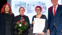 Gruppenfoto von der Urkundenverleihung mit drei Frauen, eine davon mit Urkunde, eine mit Blumenstrauß und einem Mann