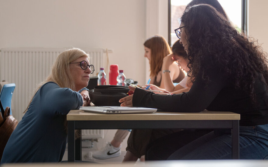 Professorin Barbara Sprick sitzt in der Hocke und spricht mit einer Studentin die am Tisch sitzt