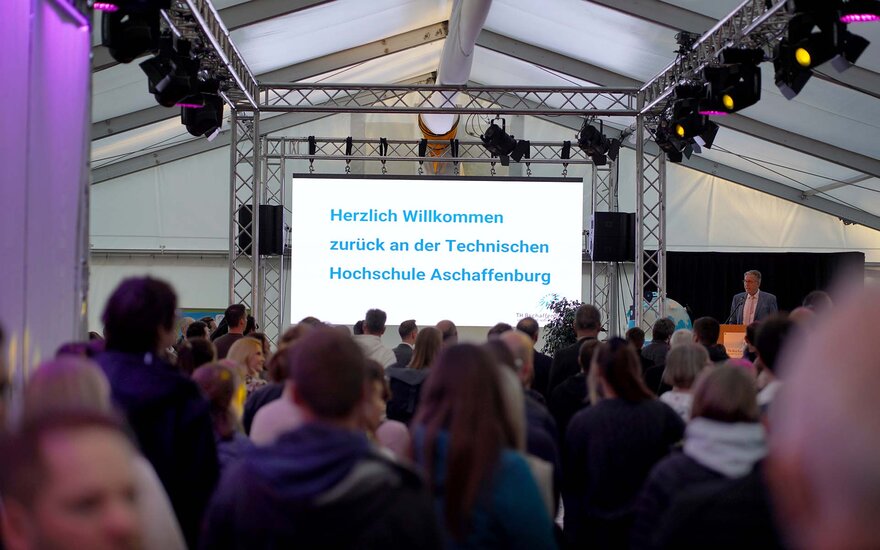 Große Gruppe von Personen im Festzelt von hinten mit Blick auf die weiße Leinwand mit der blauen Aufschrift "Herzlich willkommen zurück an der Technischen Hochschule Aschaffenburg"