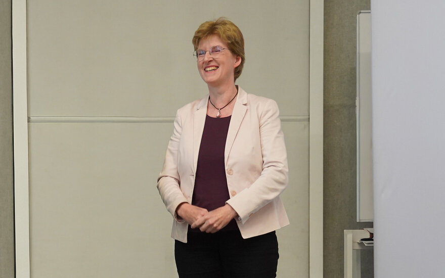 Frau mit Brille, kurzen blonden Haaren steht lächelnd mit gefalteten Händen auf der Bühne