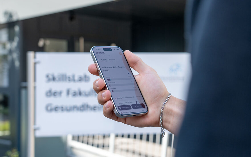 Ein Mann hält ein Handy vor einem Schild mit der Aufschrift "SkillsLab der Fakultät Gesundheitswissenschaften"