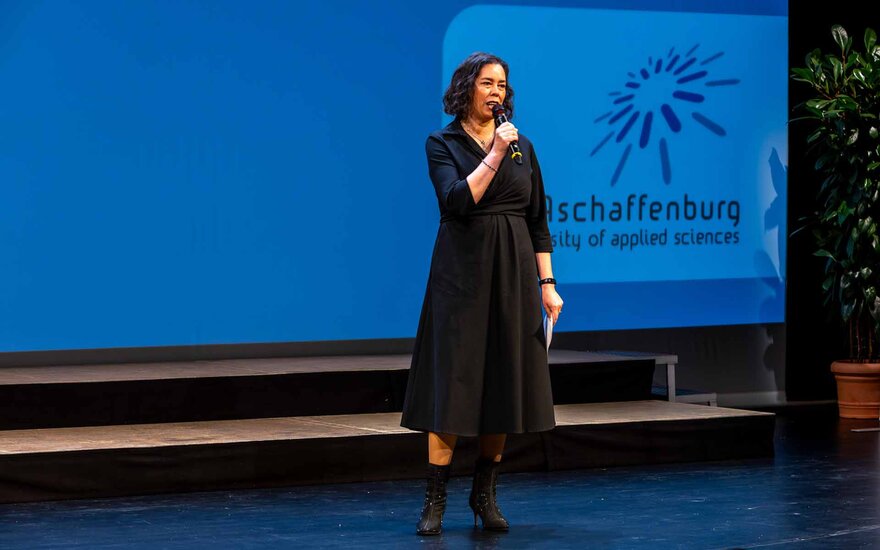 Dunkelhaarige Frau im schwarzen Kleid spricht mit Mikrophon auf der Bühne