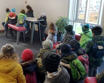 Kinder sitzen gemeinsam in einem Raum der Hochschule mit dem Rücken zur Kamera