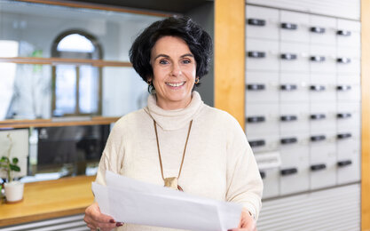 Lächelnde Frau steht vor grauen Postfächern und hat Zettel in den Händen.