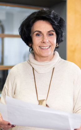 Lächelnde Frau steht vor grauen Postfächern und hat Zettel in den Händen.