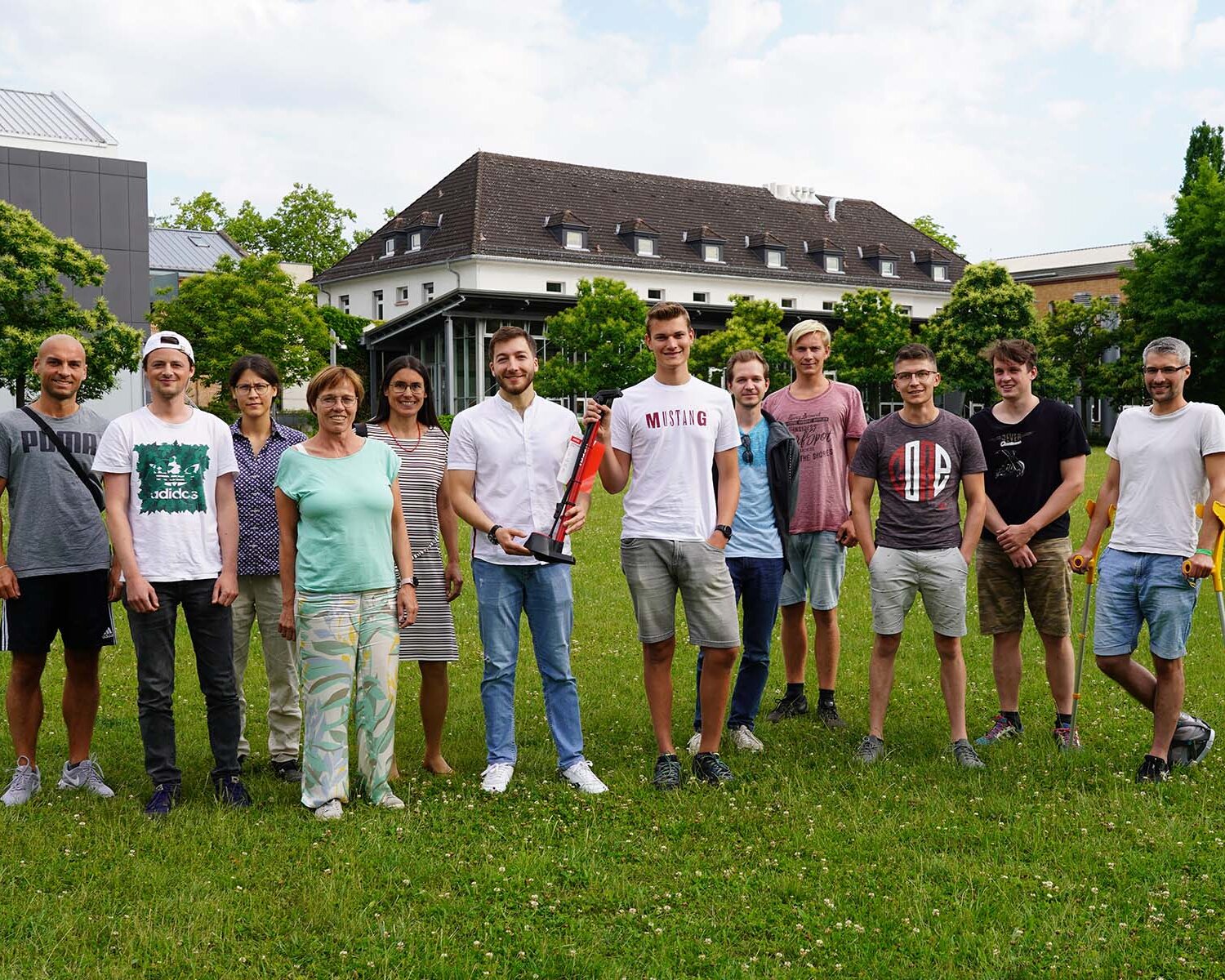 Gruppe von zwölf Personen steht auf einer grünen Wiese vor einem Gebäude und grünen Bäumen