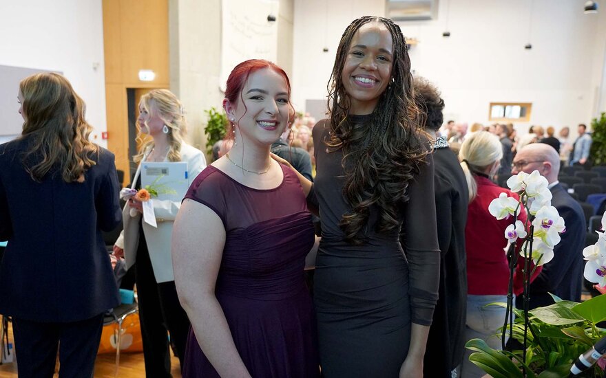 Zwei Absolventinnen stehen Arm in Arm lächelnd auf der Bühne mit den Anwesenden der Feier im Hintergrund