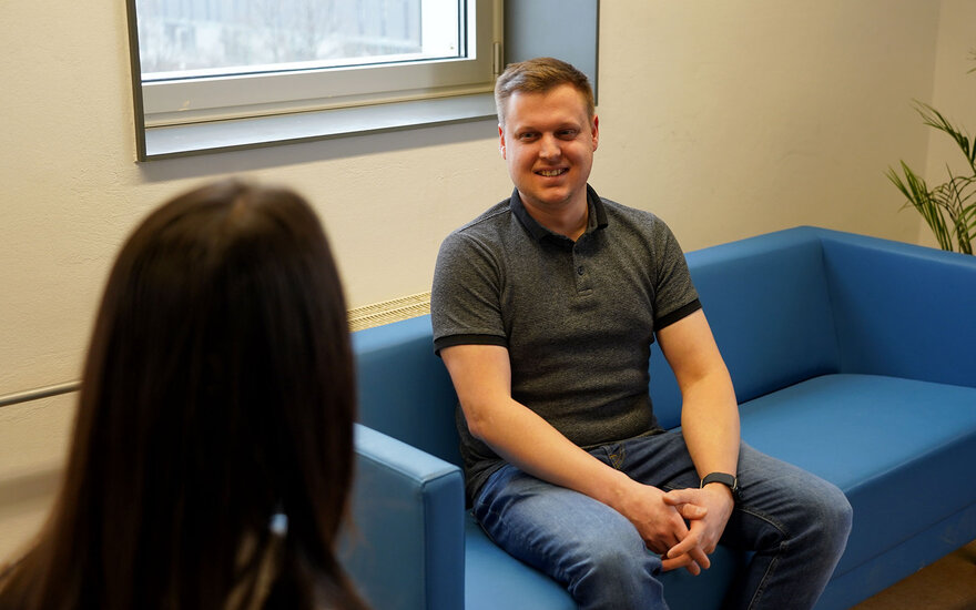 Luca Spielmann sitzt auf einer blauen Couch und redet mit einer Frau, die nur im Rückenprofil sichtbar ist.