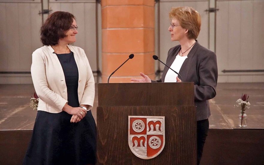 dunkelhaarige Frau und blonde Frau stehen einander zugewandt am Rednerpult