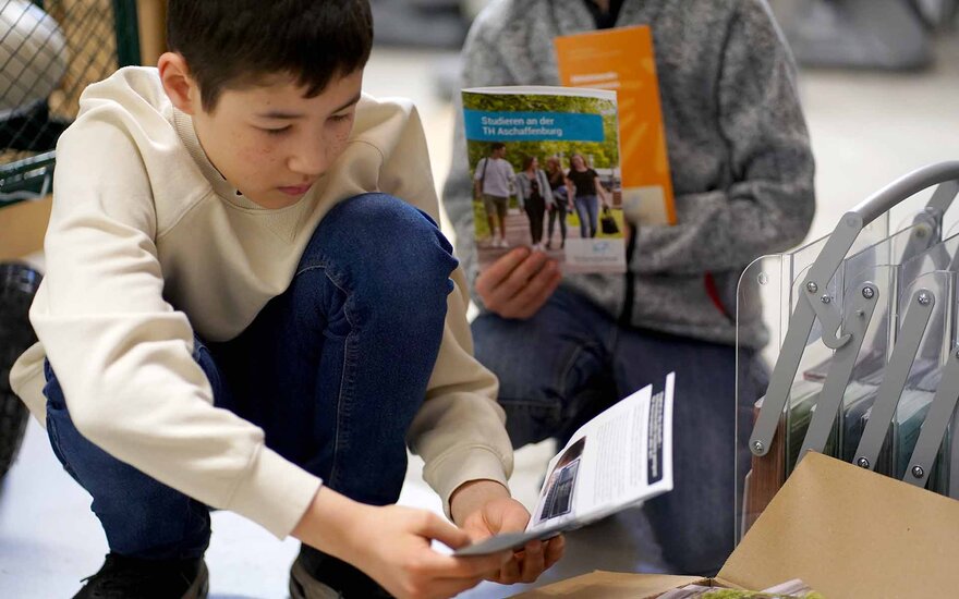 Schwarzhaariger Junge schaut hockend einen Flyer an, weiterer Junge mit Broschüre im Hintergrund