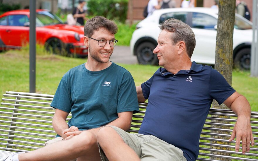 Vater und Sohn sitzen lachend nebeneinander auf einer Bank am Campus