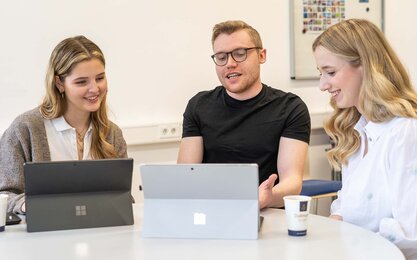 Luisa und zwei ihrer Kommilitonen sitzen vor zwei Laptops und unterhalten sich dabei lachend.