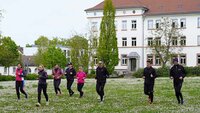 Acht Personen laufen über die grüne Campuswiese mit dem weißen Hochschulgebäude mit rotem Dach im Hintergrund