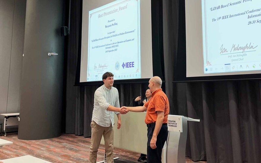 Zwei Männer geben sich die Hand. Sie stehen vor einer großen Leinwand, auf der ein Bild der Urkunde vom Best Presentation Award für Benjamin Serfling abgebildet ist. 