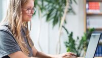 Blonde Frau sitzt an einem Tisch und arbeitet an einem schwarzen Laptop. Im Hintergund ist eine Pflanze und ein Bücherregal.