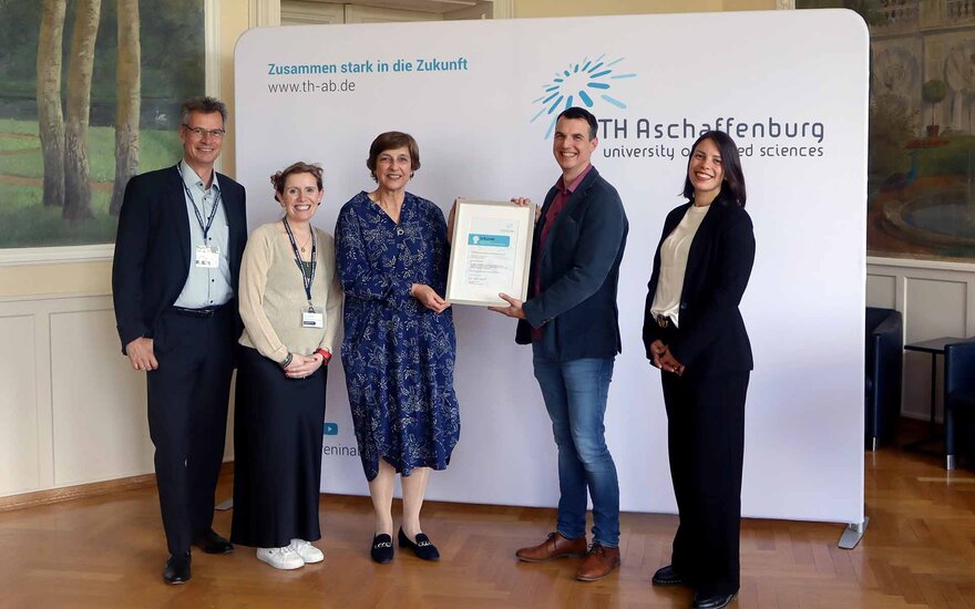 Gruppenfoto vor TH-Logo-Wand im Hocksaal mit zwei Männern und drei Frauen