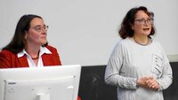 Eine Frau spricht zum Publikum, eine weitere Frau steht hinter einem Bildschirm daneben ebenfalls mit Blick zum Publikum