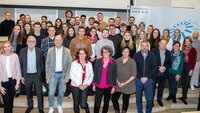 Fördernde, Stipendiatinnen und Stipendiaten stehen in der Aula der Hochschule in mehreren Reihen nebeneinander auf der Bühne, im Hintergrund ist ein Teil des blauen TH-Logos zu sehen