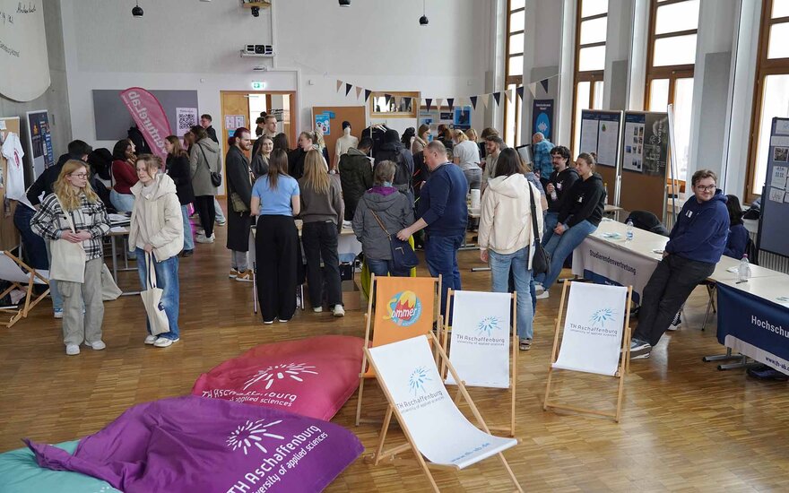 Mit Menschen gefüllte Aula mit zahlreichen Ständen sowie Sitzsäcken und Liegestühlen im Vordergrund
