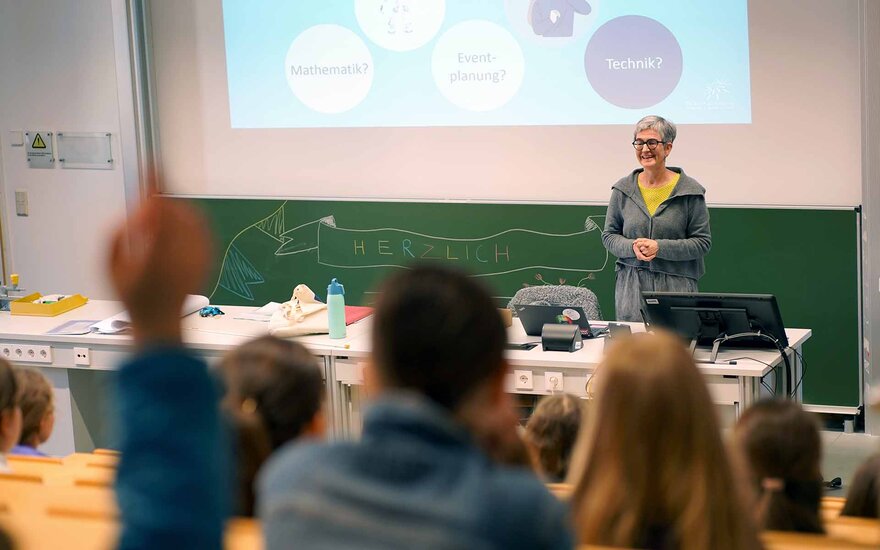 Frau steht an einem Tisch vor der Tafel und einer Leinwand im Hörsaal und lächelt die sitzenden Mädchen an.
