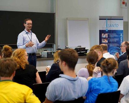 Projektleiter Professor Mewes spricht zum sitzenden Publikum im Hörsaal vor einer Tafel mit einem blau-weißen SANTO-Rollup rechts im Hintergrund