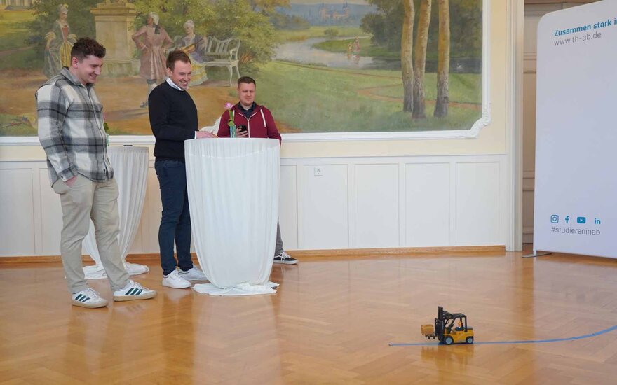 Timm steuert mit Fernbedienung einen Mini-Gabelstapler, Manuel und Luca stehen neben ihm an einem Stehtisch