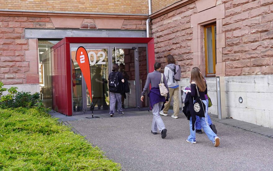 Fünf Schülerinnen und Schüler laufen auf Eingangstür von TH-Gebäude an Sparkassen-Dropflag vorbei