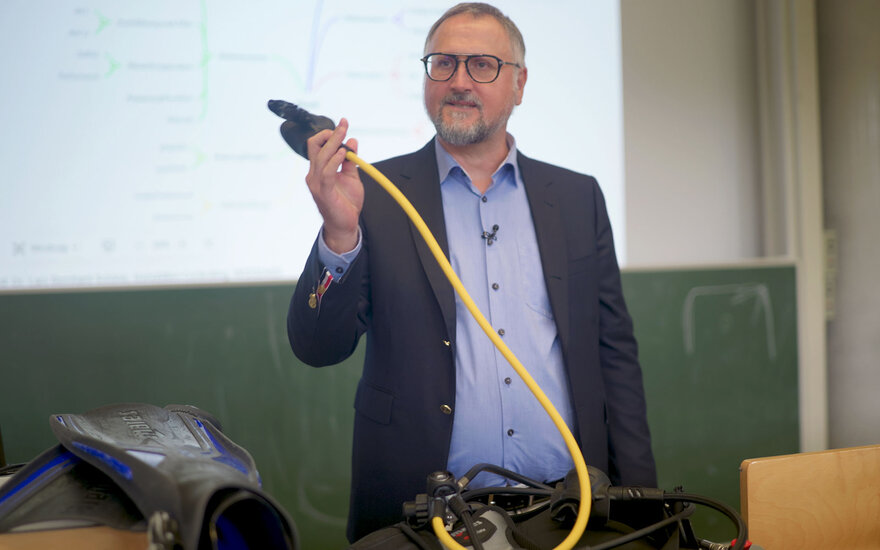Professor Lars Schöne hält den gelben Schlauch seines Tauchanzugs und zeigt ihn im Vorlesungssaal