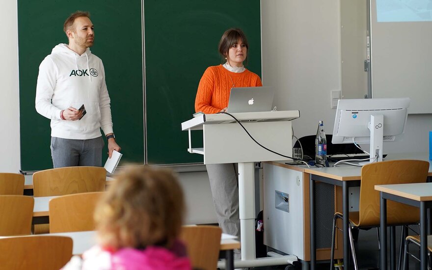Eine Frau und ein Mann stehen vor der Tafel eines Vorlesungsraums und halten eine Präsenatation am Gesundheitstag.