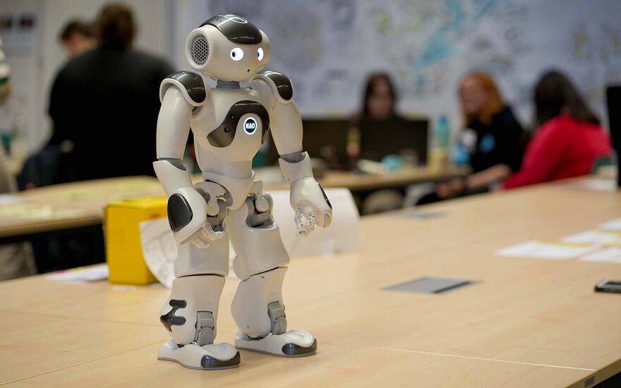 Roboter steht auf Tisch im Hintergrund sind unscharf miteinander arbeitend Mädchen zu sehen