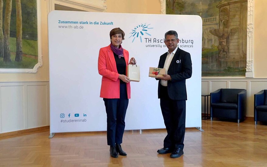 Frau in rotem Blazer und Mann im schwarzen Anzug halten je ein Geschenk in der Hand