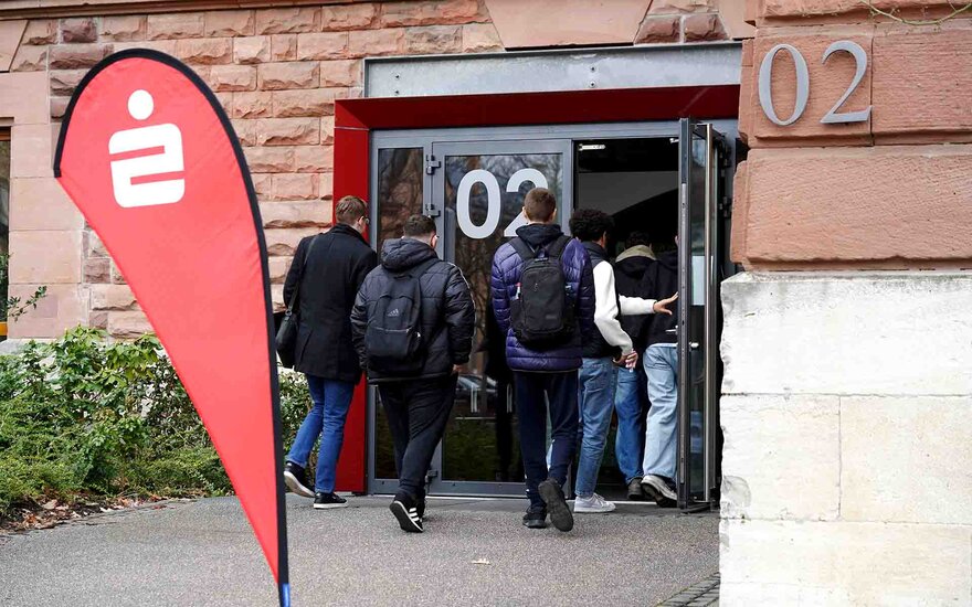 Gruppe von Jugendlichen geht durch die Eingangstür in Gebäude 02, rote Sparkassen-Beachflag steht daneben