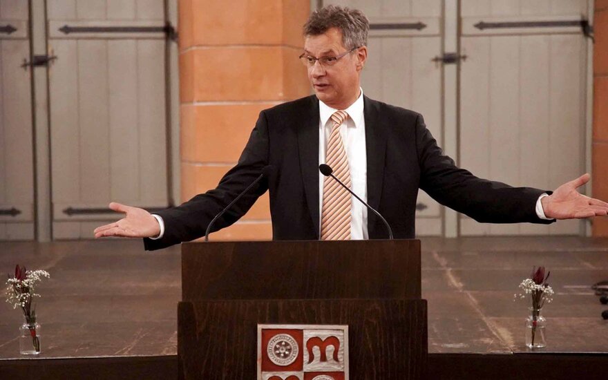 Mann mit grauen Haaren und Brille steht mit ausgebreiteten Armen am Rednerpult