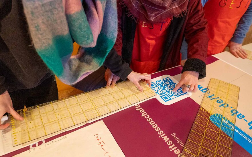 Drei anonyme Mitglieder des Gesta e.V. befinden sich an einer Werkbank und messen ein altes, lilarotes Werbebanner der TH Aschaffenburg mit großen Linealen aus. Zwei der Personen tragen rote, bestickte "Gesta" T-shirts. Die Person in der Mitte zweigt auf einem QR-Code auf dem Plakat.
