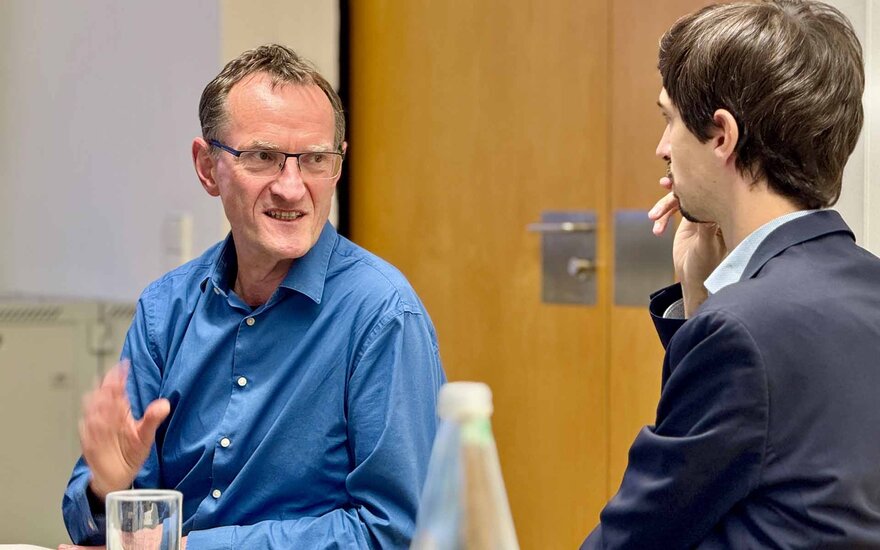 Zwei einander zugewandte Männer im Gespräch. Ein Mann im blauen Hemd ist von vorne, der andere, im schwarzen Sakko, von hinten zu sehen. 
