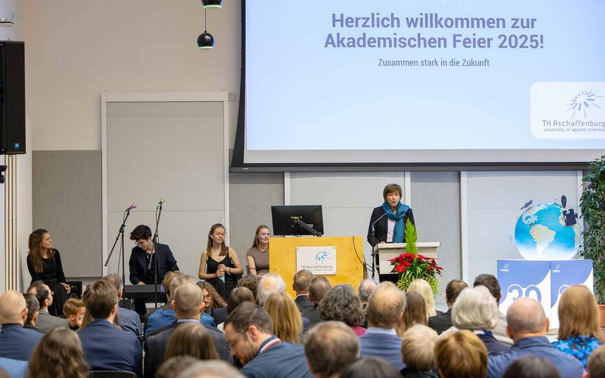 Publikum von hinten mit Blick auf Präsidentin am Rednerpult, Herzlich Willkommen auf Leinwand