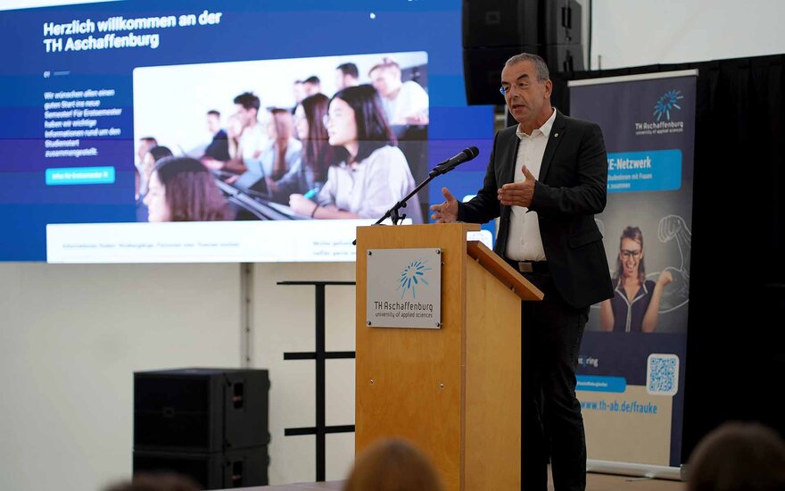 Mann mit Brille und schwarzem Anzug am Rednerpult vor Leinwand