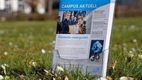 Hochschulmagazine im Prospektständer auf Wiese mit Gänseblümchen vor Gebäuden der TH